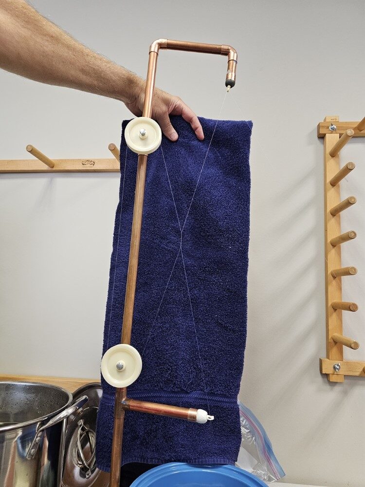A stand made of copper pipe with two pulleys up the side and an eye on a pair of arms - one at the top and one at the bottom. A dark towel is held up behind to show the path of the filament, kind of a big X connecting the pulleys and the eyes