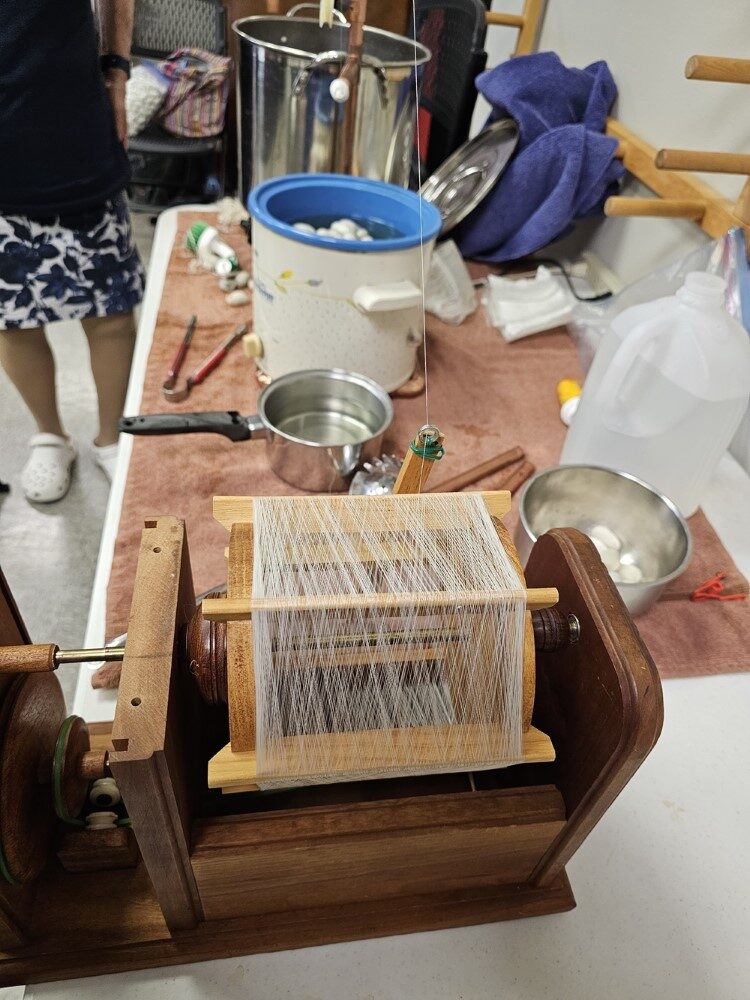 In the foreground, a wooden crank box with a Japanese-style silk reel mounted in it. In the background, various pots and tools, including a Crock Pot.