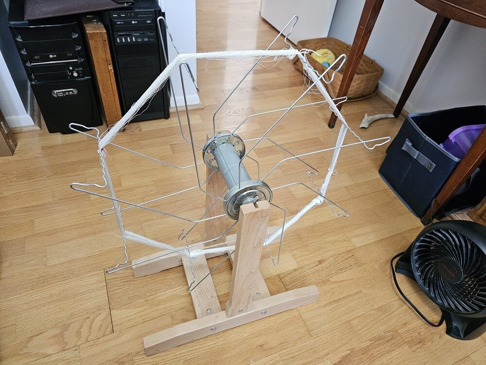A vertical skein winder, looks vaguely like a ferris wheel, with a skein of white thread connecting the arms. In the background, other junk, like old desktop computers and dog toys