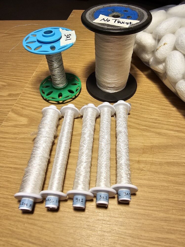 The finished yarns - five out of six of the Slick Silks for the workshop. The spool labeled 'No Twist' is filament straight from the cocoons. The spool labeled '3x1' is three filaments that have been twisted together using a spinning wheel, to make a single 'ply'. In the foreground, five white weaving bobbins, the end of each labeled with blue washi tape, like "3x2" for 3 filaments, 2 plies. In the background, a small spool with a little thread wond on it, and a larger spool half-full of untwisted filament.